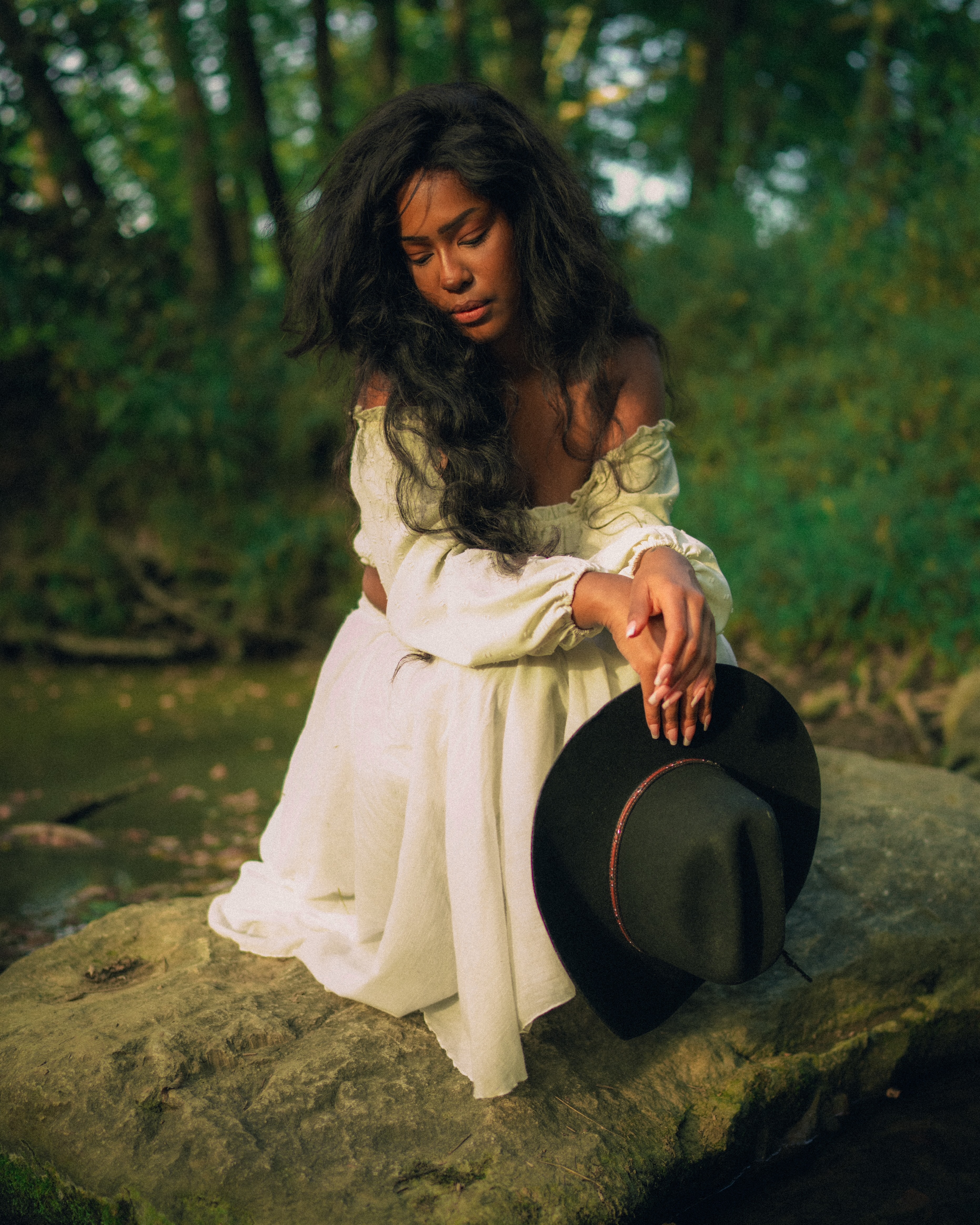 Woman with tousled dark hair looking down, seated on a stone ledge in a white off-shoulder dress, dangling a black hat from one hand over a wooded creek backdrop.