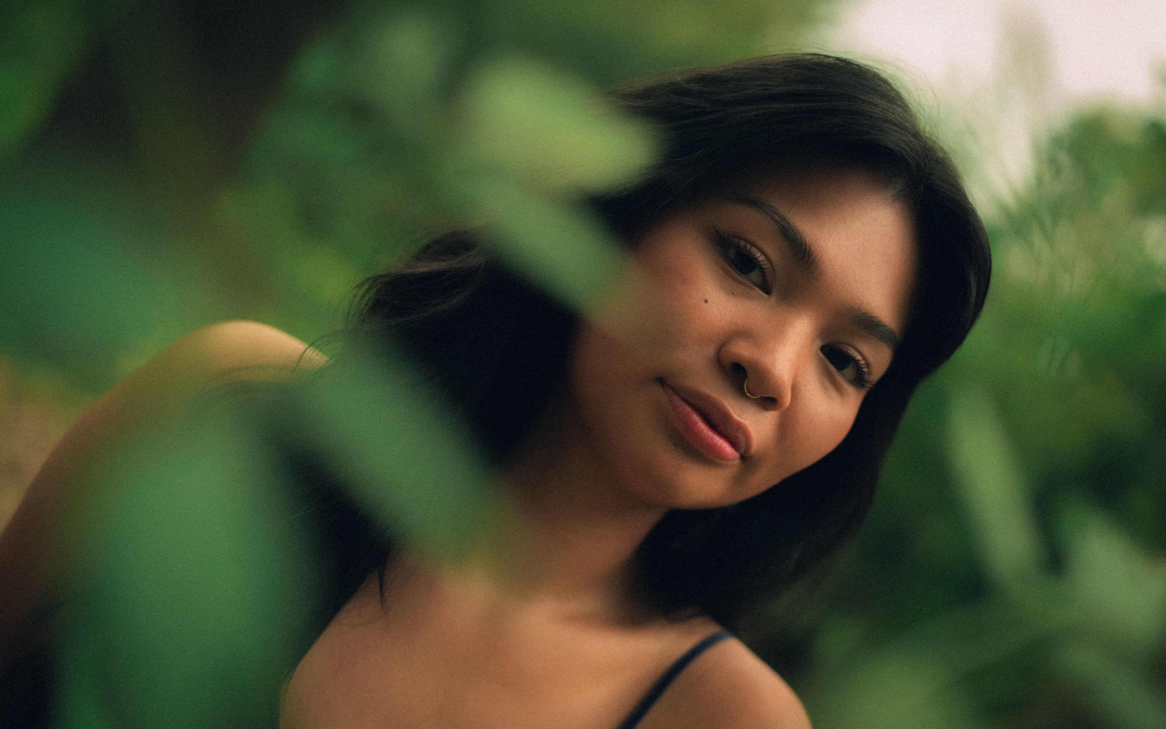 Woman with a small nose ring and beauty mark tilts her chin down, gazing through a gap in blurred green foliage, wearing a thin dark strap top.