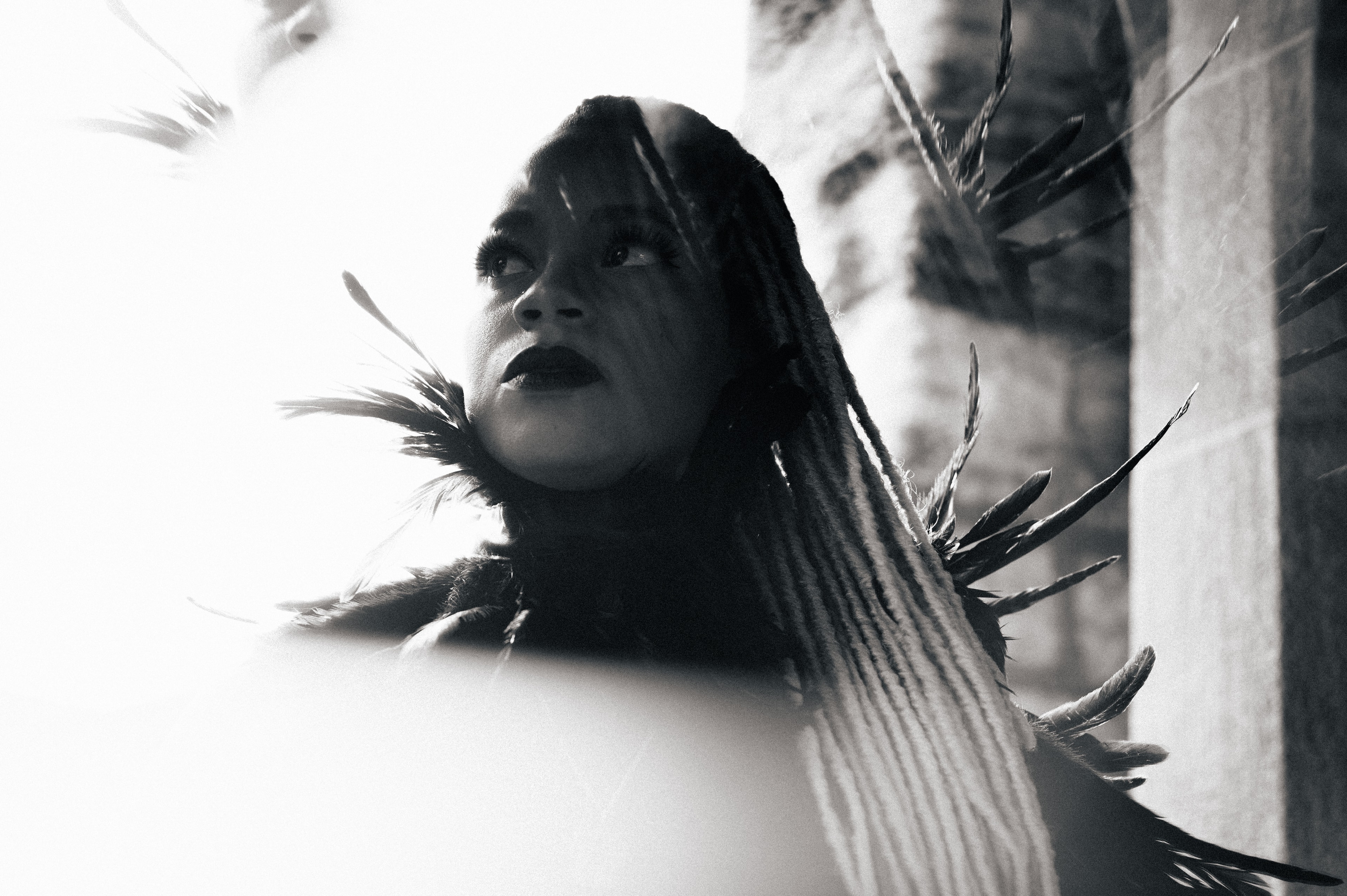 Black-and-white low-angle portrait of a woman with long striped braids and a spiky feathered collar, chin tilted up, half her face lit and half in shadow beside a stone wall.