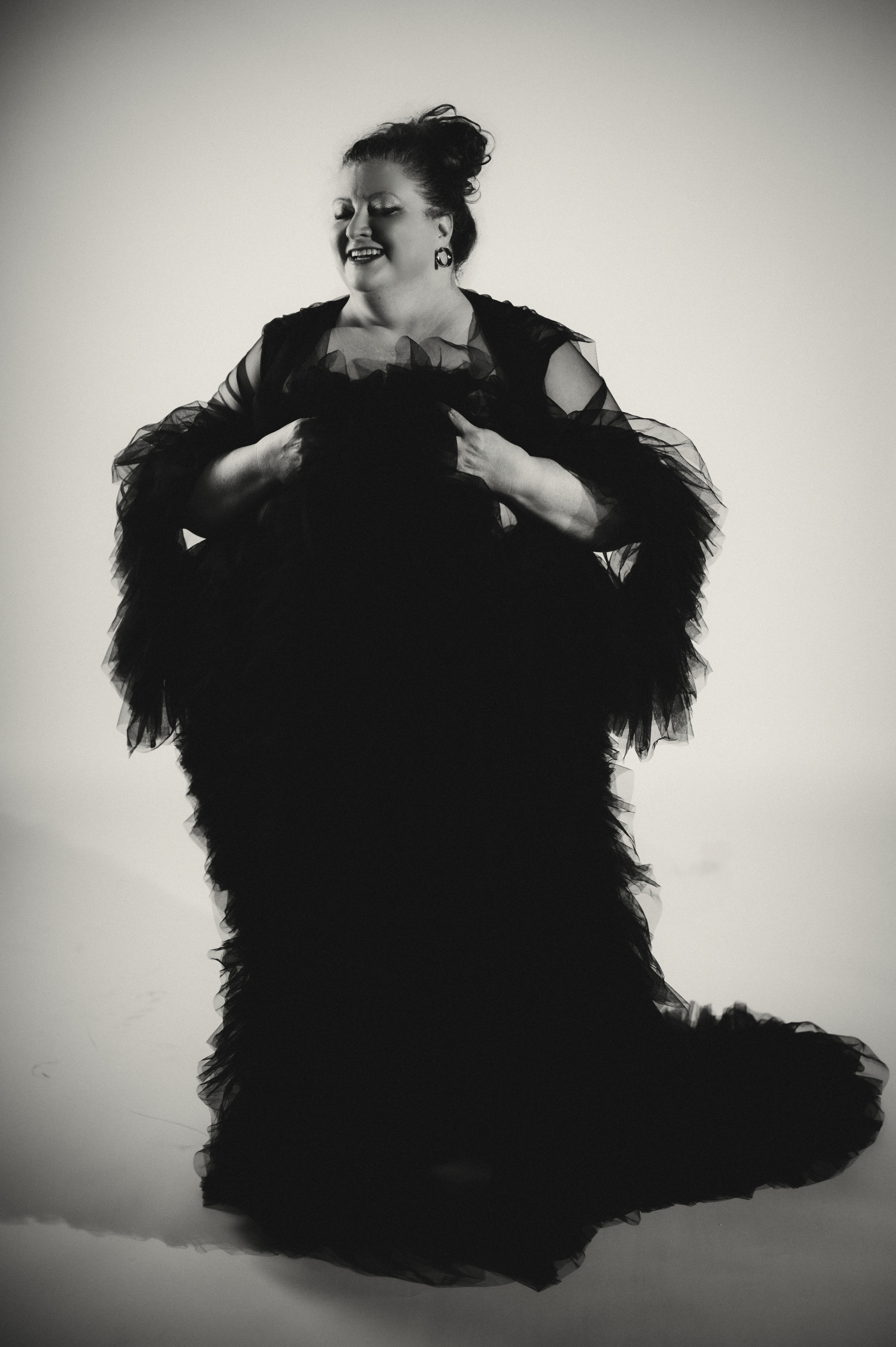 Black-and-white studio portrait of a woman with a loose top-knot and dangling round earrings, eyes closed and smiling as she clutches layers of black tulle ruffles at her chest, the full gown cascading into a long train on the floor.