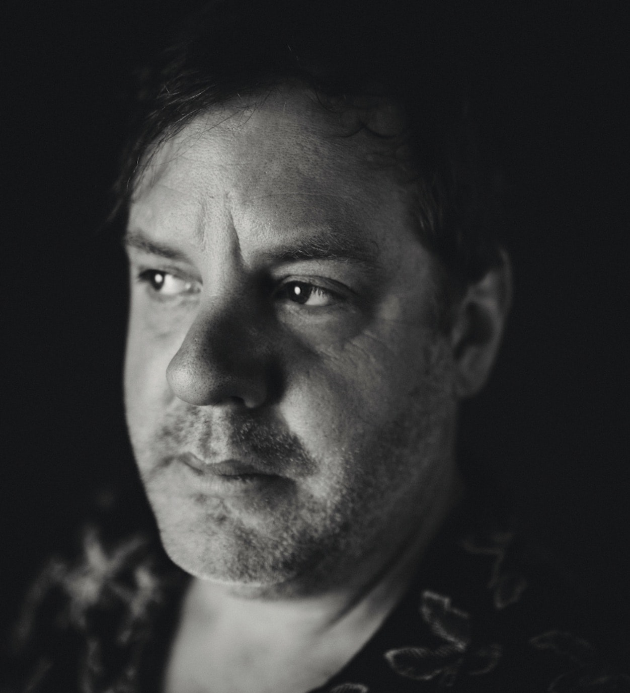 Close-up black-and-white portrait of a heavyset man in a patterned shirt, furrowed brow, eyes cast to the side, deep shadow engulfing half his face.