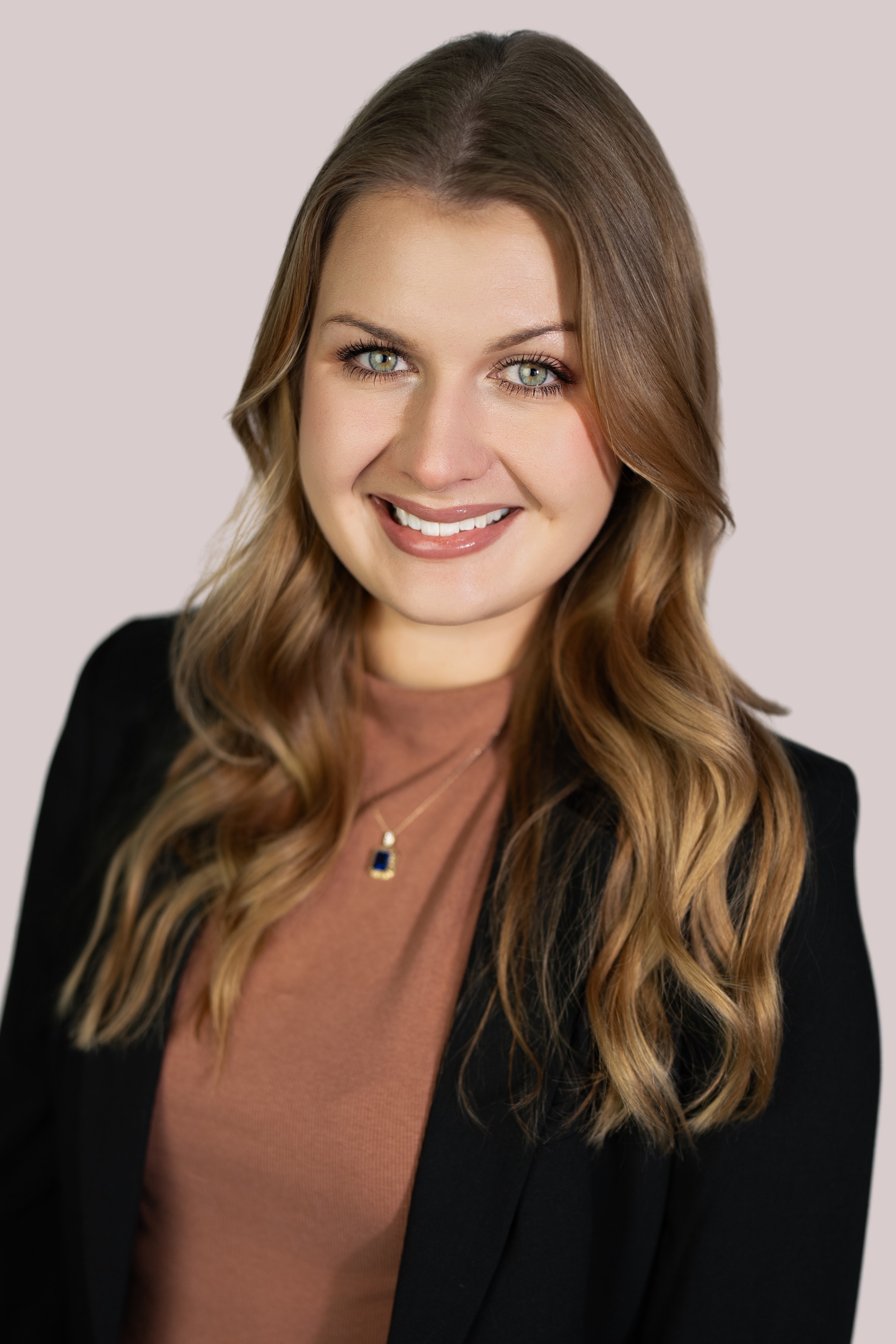 Woman with long wavy caramel-highlighted hair centered part, green eyes, glossy nude lipstick, smiling directly at camera in black blazer, rust top, and gold sapphire pendant against a blush-pink backdrop.