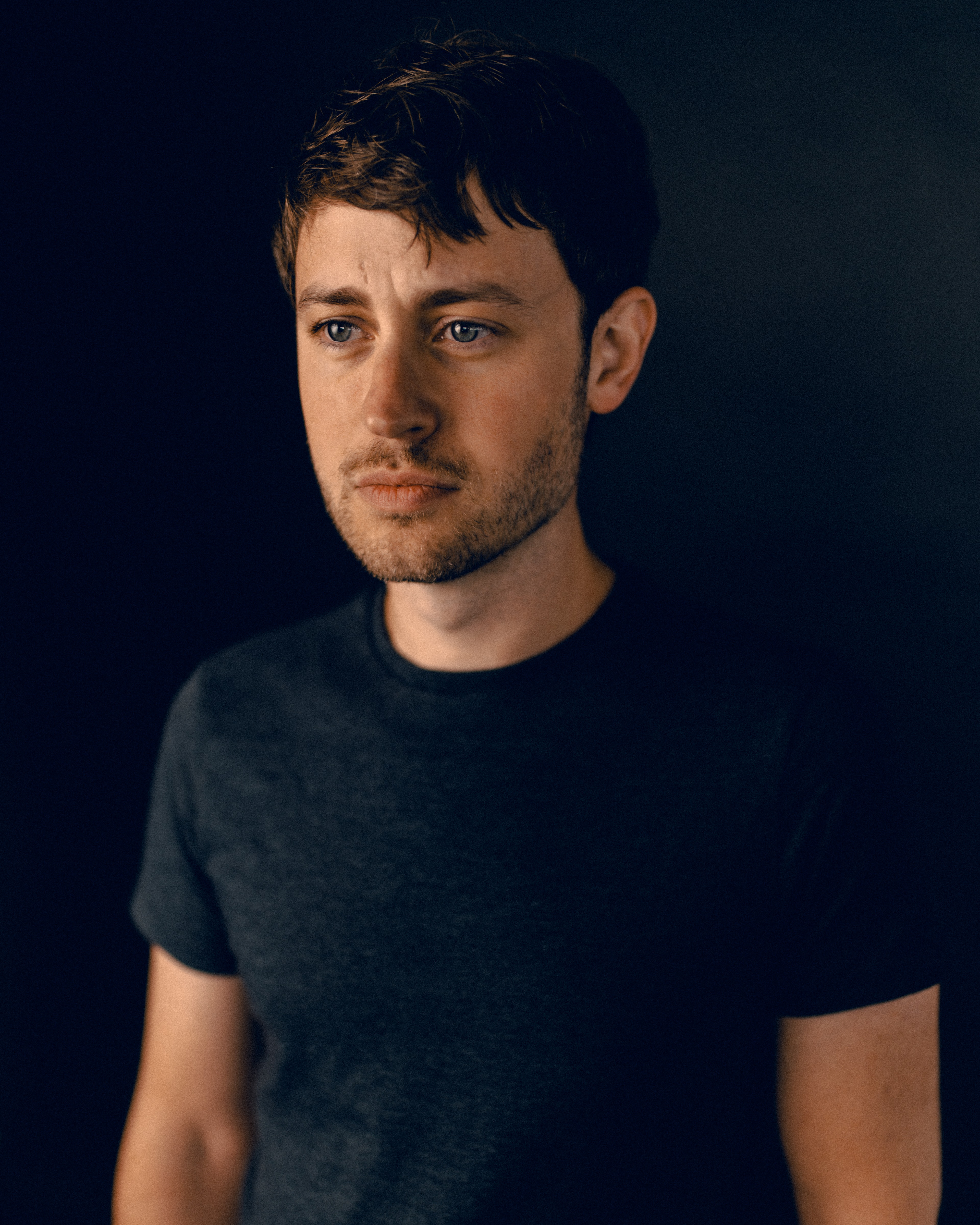 Man with tousled brown hair and light stubble looks off to the right, face lit warmly from below-left, dark t-shirt fading into black background.