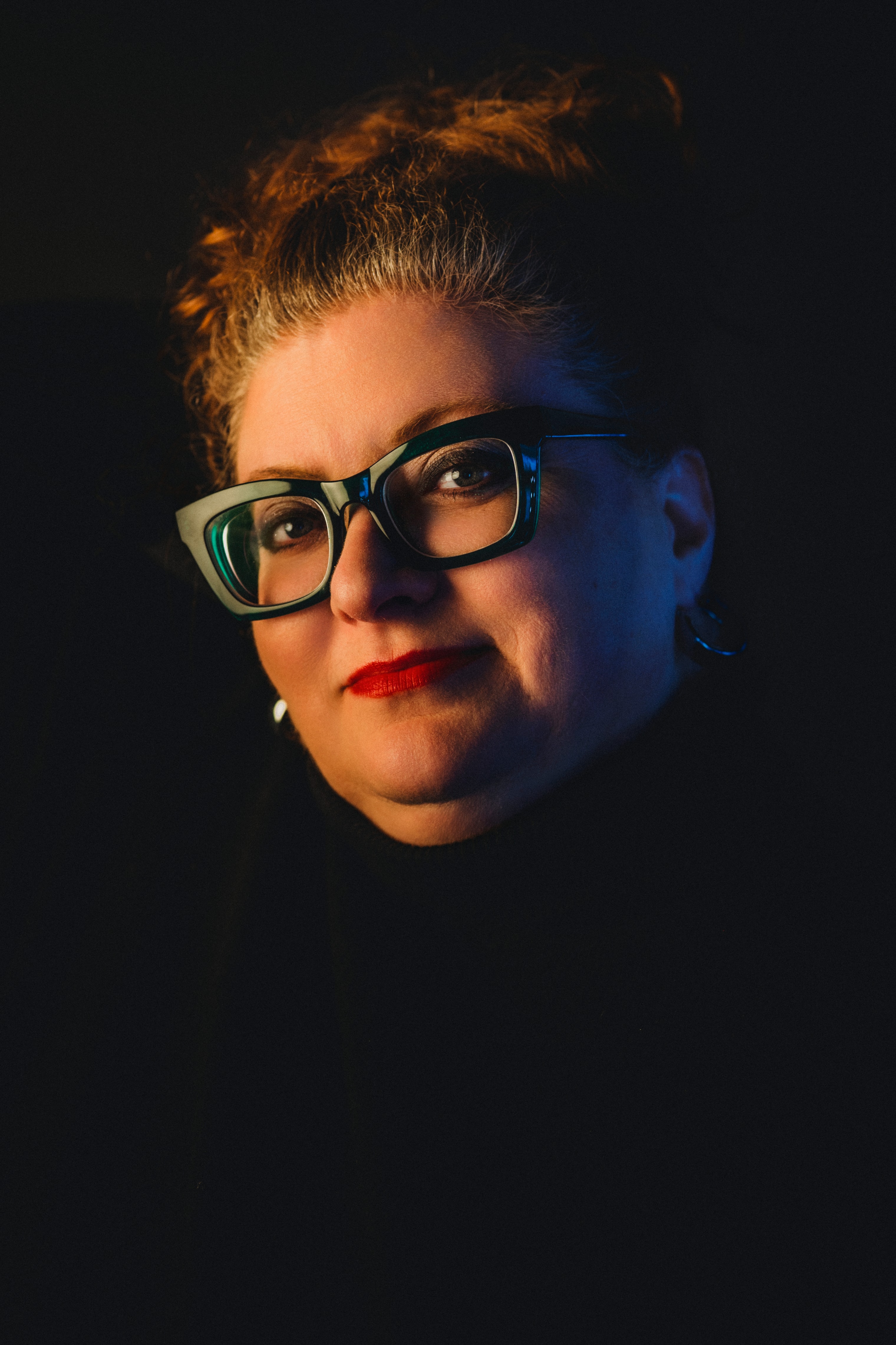 Woman with curly hair swept up, teal-framed cat-eye glasses, and red lipstick gazes upward as amber light warms her face and blue light rims her ear and earring against total black.