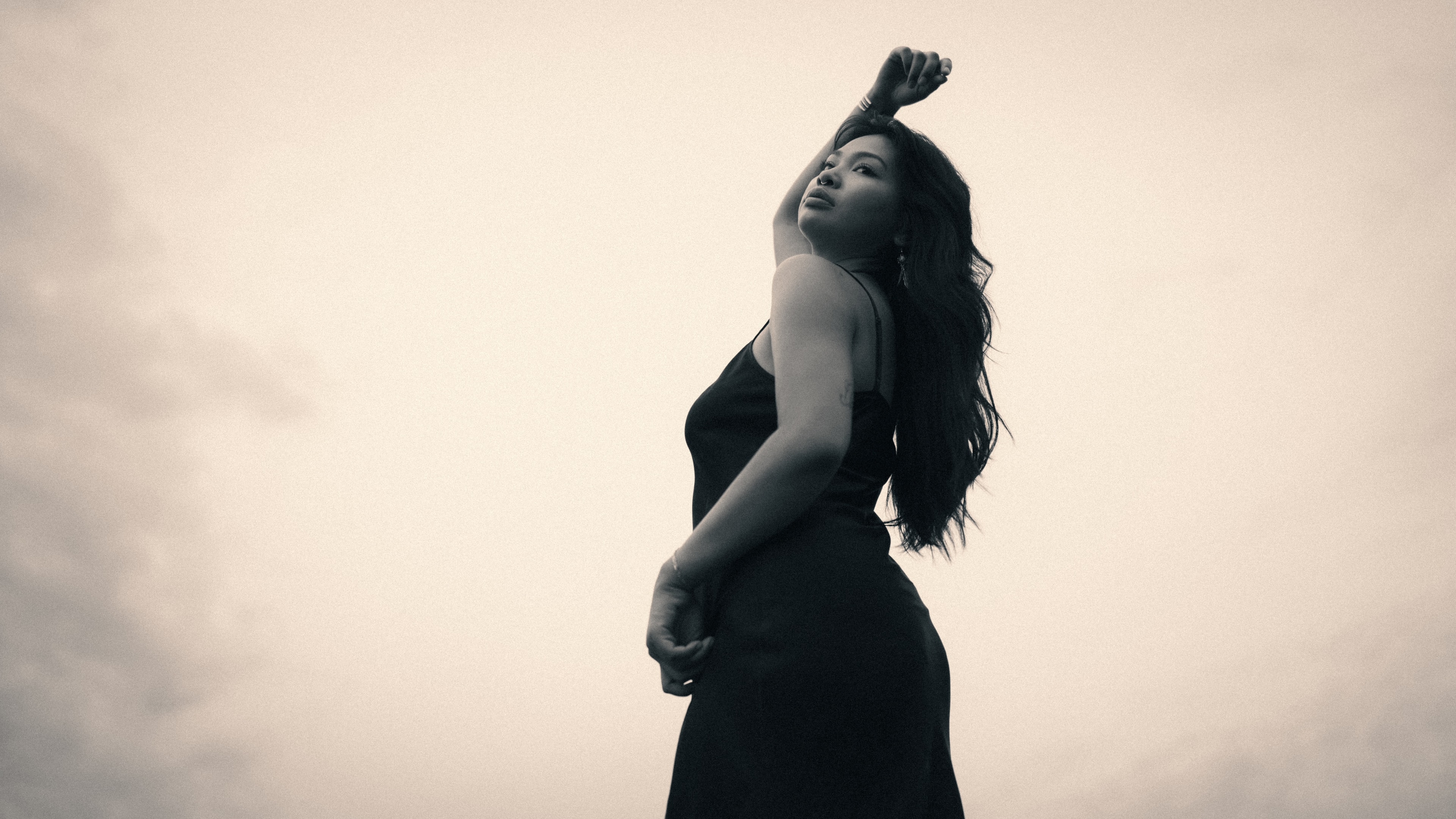 Low-angle silhouette of a woman in a black slip dress, head tilted back and one fist raised above her hair, against a pale hazy sky in this Columbia, Missouri portrait.